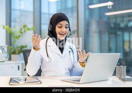 Une femme médecin, portant un hijab et un casque, s'engage dans une consultation de télésanté via un ordinateur portable, faisant des gestes avec excitation. Banque D'Images