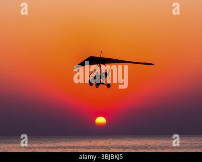 La personne volant sur un tricycle avec le moteur contre le deltaplane vole contre le coucher du soleil Banque D'Images