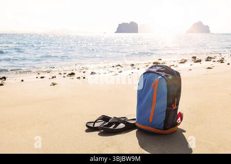 Sac à dos avec tongs sur une belle plage de sable contre le soleil. Concept de voyage minimaliste Banque D'Images