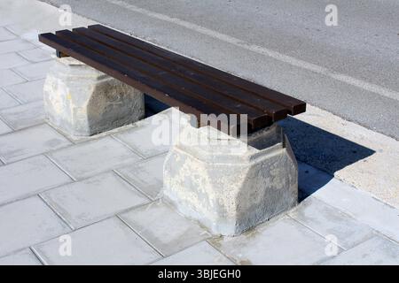Banc de parc simple avec des lattes en bois sombre et des supports en béton robustes se dresse sur un trottoir pavé près de la route, projetant une ombre en plein soleil, off Banque D'Images