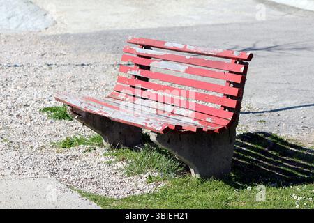 Ancien banc de parc rouge avec peinture pelable et supports en béton se trouve sur le gravier et l'herbe dans un espace public extérieur, montrant des signes d'âge et d'utilisation dans Bright Banque D'Images