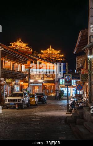 Shangri-la, Chine - 20 octobre 2024 : des bâtiments illuminés et des voitures garées bordent une charmante rue dans la ville antique de Dukezong la nuit. Banque D'Images