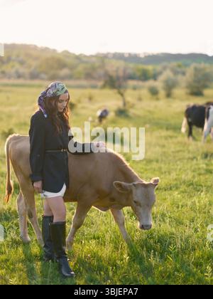 Jeune femme élevant des vaches sur un pâturage vert dans la campagne. Scène extérieure d'été rural avec le bétail, la nature et la vie agricole contre belle l Banque D'Images