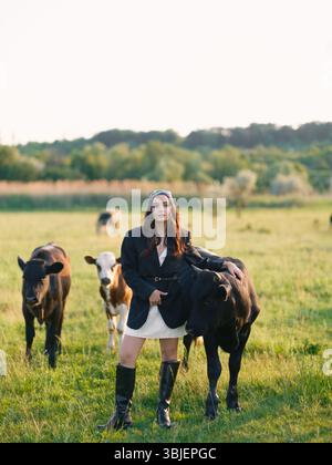Jeune femme élevant des vaches sur un pâturage vert dans la campagne. Scène extérieure d'été rural avec le bétail, la nature et la vie agricole contre belle l Banque D'Images