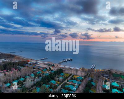 Vue aérienne du lac Issyk Kul sous un ciel couvert de coucher de soleil, montrant une station balnéaire avec des bâtiments nichés parmi les arbres et les eaux calmes du lac Banque D'Images