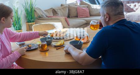 Matin en famille avec thé : fille et homme assis à une table en bois avec citron, viennoiseries et théière dans un salon lumineux et confortable Banque D'Images