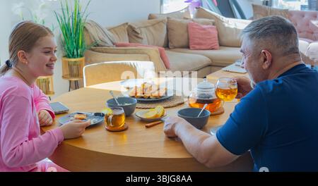 Matin en famille avec thé : fille et homme assis à une table en bois avec citron, viennoiseries et théière dans un salon lumineux et confortable Banque D'Images