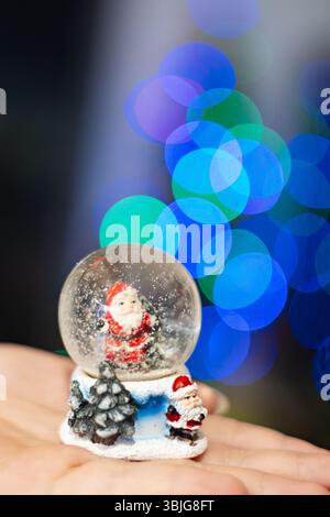 Gros plan extrême d'une boule de neige festive avec la figurine du Père Noël tenue à la main. La neige étincelante et le bokeh bleu-vert vif créent une sensation magique de vacances d'hiver Banque D'Images