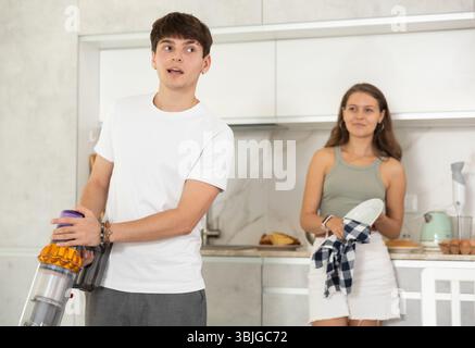 Guy aspirateur dans la cuisine tout en communiquant avec sa petite amie sécher la vaisselle avec une serviette Banque D'Images