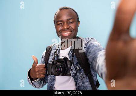 Vlogger masculin afro-américain donnant les pouces à son public en ligne, s'enregistrant lui-même en studio. POV de photographe noir joyeux faisant un geste d'accord vers l'appareil photo. Banque D'Images