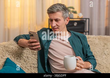 Jeune homme à la maison commence le matin assis sur un canapé avec du thé, lisant smartphone avec une concentration calme. Le gars caucasien semble paisible, concentré sur les nouvelles et détendu, appréciant une boisson chaude dans un moment calme à la maison. Banque D'Images