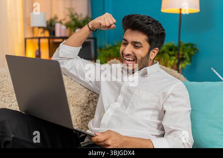 Homme indien célébrant le succès à la maison, acclamant avec joie, réagissant émotionnellement à l'écran d'ordinateur portable. Mec arabe assis sur le canapé, se sentant heureux, excité, satisfait après la réalisation du projet ou l'affaire Banque D'Images