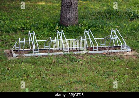 Un porte-vélos en métal blanc avec plusieurs fentes se trouve vide sur une pelouse herbeuse dans un parc, entouré de feuillage vert et ombragé par un arbre, prêt à l'emploi b Banque D'Images