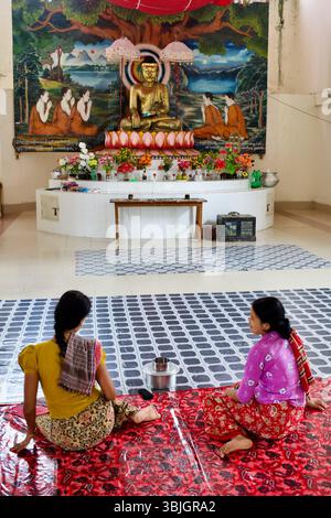 Des femmes assises en prière au temple bouddhiste Srimangal à Kuakata, Bangladesh, face à une murale vive et à Bouddha doré assis. Banque D'Images