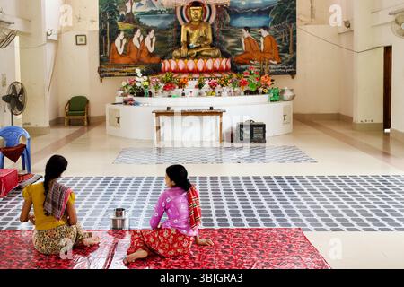 Des femmes assises en prière au temple bouddhiste Srimangal à Kuakata, Bangladesh, face à une murale vive et à Bouddha doré assis. Banque D'Images