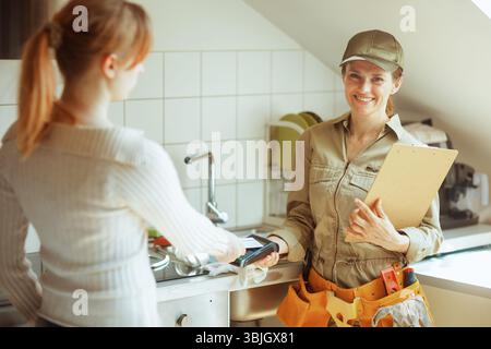 Un plombier souriant accepte un paiement par carte d'un client dans un cadre lumineux de cuisine. La scène transmet un service de réparation à domicile efficace et professionnel. Banque D'Images