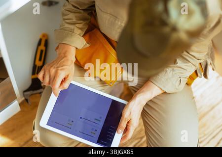 Une femme plombière examine les tâches sur une tablette assise avec des outils à proximité, organisant les prochaines étapes pour résoudre un problème de plomberie de cuisine. Banque D'Images