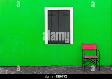 Une chaise solitaire se dresse contre un mur vert éclatant avec une fenêtre à cadre blanc avec des volets sombres fermés à Burano, en Italie. Banque D'Images