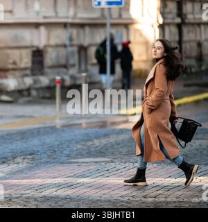 Avec ses cheveux coulant dans la brise, une jeune femme marche gracieusement sur des pavés, mettant en valeur son manteau à la mode et sa tenue décontractée dans un urb animé Banque D'Images