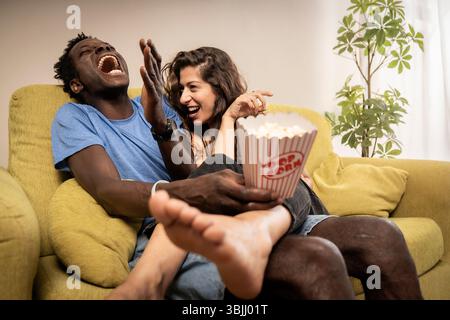 Un jeune couple exubérant et diversifié éclate de rire en regardant un film de comédie et en mangeant du pop-corn à la maison. Un moment authentique et joyeux de sh Banque D'Images