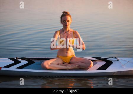 Femme calme faisant de la méditation yoga sur paddleboard dans la lumière tôt le matin, en se concentrant sur la pleine conscience, l'équilibre de l'eau et la tranquillité spirituelle. Banque D'Images