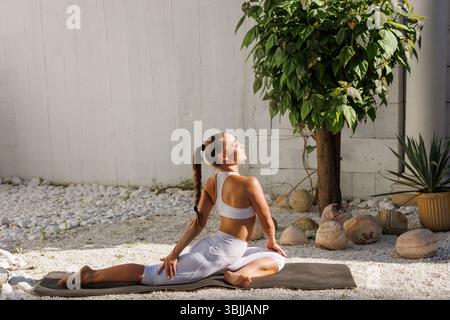 Femme en vêtements d'action blancs pratiquant une torsion de yoga assis à l'extérieur, axée sur la flexibilité, la relaxation et la pleine conscience. Banque D'Images