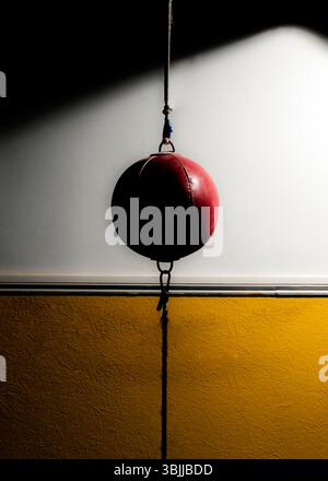 Un sac de vitesse en cuir rouge est suspendu dans une pièce sombre. Il est suspendu au plafond par une corde et une chaîne. La moitié supérieure de la pièce est blanche. Banque D'Images