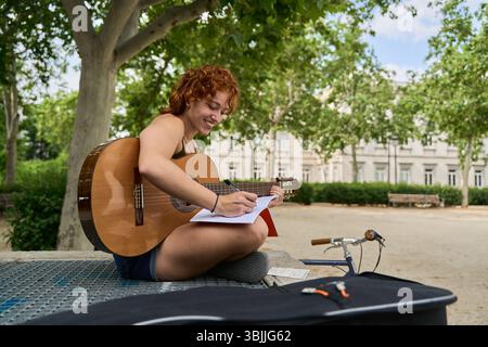 Jeune musicienne femme rousse composant une chanson avec guitare acoustique et écrivant des notes sur un bloc-notes assis dans un parc Banque D'Images