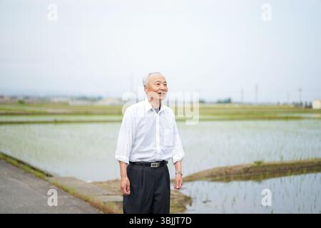 Dans les campagnes japonaises, un Japonais de la fin des années 70 marche le long d'une route agricole entourée de larges rizières. Il porte une chemise blanche et un chapeau, appréciant le fr Banque D'Images