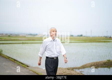 Dans les campagnes japonaises, un Japonais de la fin des années 70 marche le long d'une route agricole entourée de larges rizières. Il porte une chemise blanche et un chapeau, appréciant le fr Banque D'Images