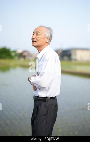 Dans les campagnes japonaises, un Japonais de la fin des années 70 marche le long d'une route agricole entourée de larges rizières. Il porte une chemise blanche et un chapeau, appréciant le fr Banque D'Images