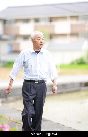 Dans les campagnes japonaises, un Japonais de la fin des années 70 marche le long d'une route agricole entourée de larges rizières. Il porte une chemise blanche et un chapeau, appréciant le fr Banque D'Images