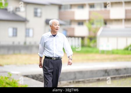Dans les campagnes japonaises, un Japonais de la fin des années 70 marche le long d'une route agricole entourée de larges rizières. Il porte une chemise blanche et un chapeau, appréciant le fr Banque D'Images