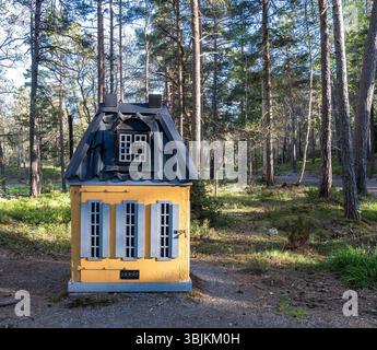 Une petite maison jaune fantaisiste comme une poubelle un toit noir et des fenêtres décoratives, situé dans une zone boisée. Les arbres environnants créent une atmosphère sereine Banque D'Images