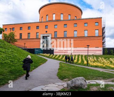 Stockholm, Suède - 11 mai 2025 : une bibliothèque orange moderne à Stockholm avec un sommet circulaire, entouré de verdure et de fleurs. Les gens marchent le long d'un p. Banque D'Images
