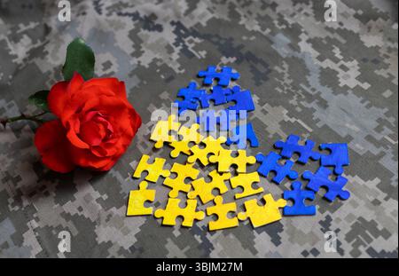 Les pièces de puzzle de couleurs bleu jaune sont pliées en cœur, fleur de rose sur uniforme de camouflage. Concept de cœur brisé par la guerre des Ukrainiens. Mémoire éternelle Banque D'Images