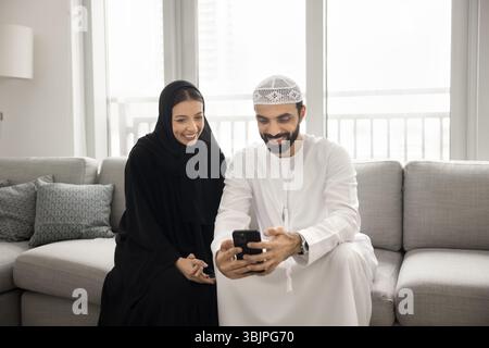Heureux couple arabe dans l'usure musulmane à l'aide d'un smartphone sur le canapé Banque D'Images