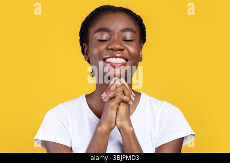Femme noire priant en tenant les mains dans le geste de prière, fond jaune Banque D'Images