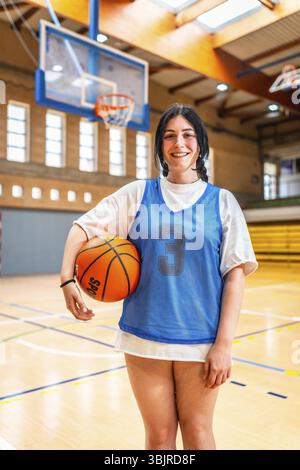 Athlète féminine souriant et tenant le basket-ball dans un gymnase, prêt pour l'entraînement ou la compétition Banque D'Images