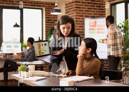 Professionnel du marketing utilisant l'Internet pour afficher des stratégies publicitaires, analysant les données des clients dans l'espace de travail moderne. Deux collègues femmes se concentrant sur l'analyse commerciale sur ordinateur portable. Banque D'Images