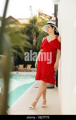Femme élégante en robe rouge et chapeau debout par cactus, oranger sous la lumière du soleil dans le jardin d'été Banque D'Images