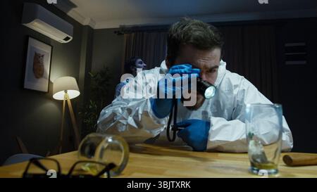 Enquêteur en costume de protection utilise une lampe de poche pour inspecter les preuves en verre sur la table dans une pièce faiblement éclairée, suggérant une analyse de scène de crime. Banque D'Images