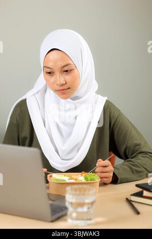 Une belle et heureuse femme musulmane asiatique mange sa salade saine tout en travaillant sur son ordinateur portable. Travail à domicile, style de vie Banque D'Images