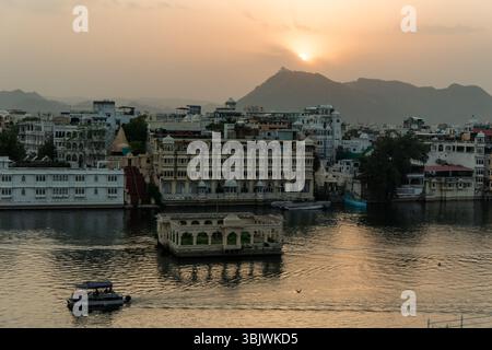 Coucher de soleil doré sur le lac Pichola illuminant Udaipur City Palace, Lake Palace Hotel et Jag Mandir avec des bateaux naviguant sur les eaux calmes et moun Banque D'Images