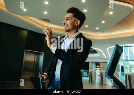 Un comptable d'entreprise fait signe de la main et accueille un collègue dans un hall élégant de bureau. Habillé en tenue professionnelle, il respire la confiance et la convivialité. Banque D'Images