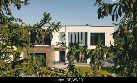 Maison moderne derrière des branches de pin avec des cônes. Une maison contemporaine de deux étages partiellement cachée derrière des branches de pin luxuriantes chargées de cônes. Banque D'Images
