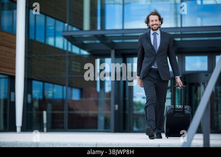 Homme descendant des marches en béton, tirant une valise roulante noire et souriant à l'entrée du bureau Banque D'Images