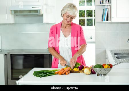 Femme âgée hachant des carottes des échalotes de brocoli sur la planche à découper de l'îlot de cuisine avec passoire Banque D'Images