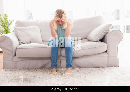 Femme blonde est assise sur un canapé gris, tenant la tête et se penchant en avant près de la plante et des fenêtres Banque D'Images