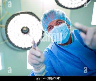 Chirurgien se penchant et effectuant une procédure tenant le scalpel sous des lumières chirurgicales vives dans le BLOC OPÉRATOIRE Banque D'Images